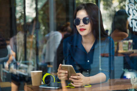 happy asian woman wearing sunglasses, holding her phone with her headphone on the table in a coffee shop, seen through window with reflectionの写真素材