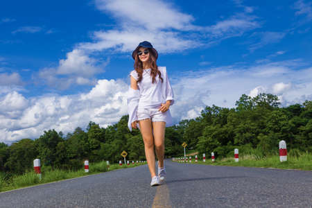 young attractive happy asian teenager woman, in white shirt, short, sunglasses and hat, walking casually and confidently on a road surrounded by nature, concept of carefree and happy free timeの写真素材