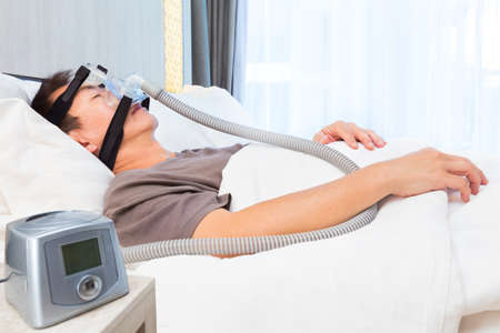 middle age asian man sleeping in his bed wearing CPAP mask connecting to air hose and CPAP machine,...