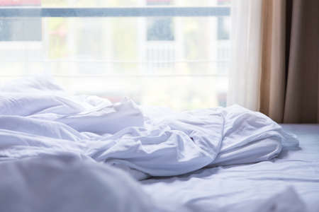 unmade bed with white sheet and blanket, bright soft morning light coming through the window,...