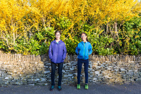 happy asian girls standing in front of old stone wall with majestic yellow flowers of Coswolds,...