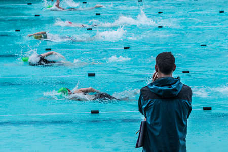 Male swimming coach standing by the swimming pool in the rain watching swimmers racing by, good for coaching or training conceptの写真素材