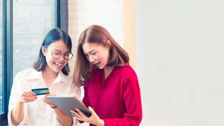 Two happy young asian women online purchasing using their credit card and computer tablet at modern...