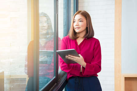 Happy asian business woman standing by large window, holding her computer tablet, looking out the window, thinking about her successful business, good for woman in business theme, room for copyspaceの写真素材