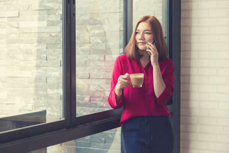 Young beautiful asian woman standing by a large window using her phone and holding a cup of coffee, vintage retro color style, room for copyspaceの写真素材