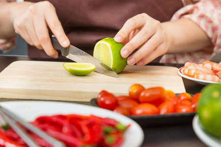 Female hands cutting green lime on wooden cutting board in her home kitchen, surrounded by other food ingredients, good for home cooking healthy conceptの写真素材