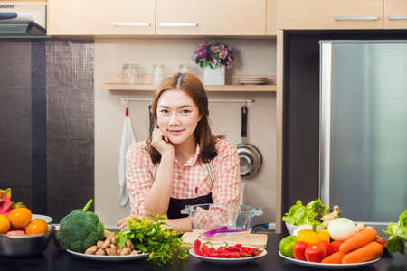 Proud attractive asian female cook standing in her home kitchen and smiling at camera, with various vegetables and fruits on the counter top, classice vintage brown color tone, good for cooking or healthy lifestyle theme conceptの写真素材