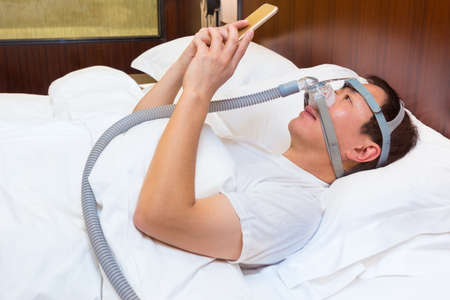 Middle age asian man laying in his bed wearing CPAP mask and using his smart phone at night