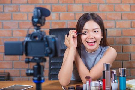 Young happy asian woman vlogging to her fan page showing how to apply mascara and put make up onの写真素材
