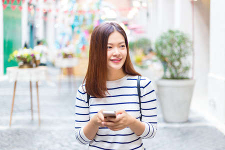Happy young attractive asian woman in casual clothes using her smart phone to connect to her friends through social media while walking outside of shopping plaza, good for casual lifestyle conceptの写真素材