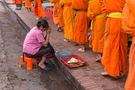LUANG PRABANG, LAOS - 29 June 2018 - Old woman prays while buddhist monks walk by in Luang Prabang, Laos on June 29, 2018のeditorial素材