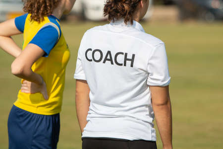 Back of sport coach wearing COACH shirt at an outdoor sport field, standing with a soccer player, watching the gameの写真素材