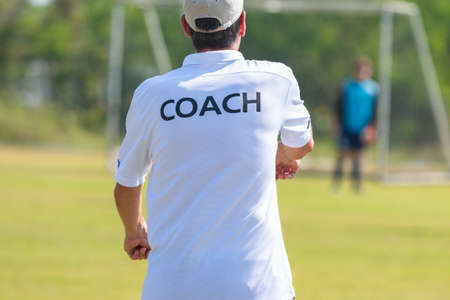 Back of football coach wearing white COACH shirt at an outdoor sport field coaching his goalie player during a game, good for sport or coaching conceptの写真素材