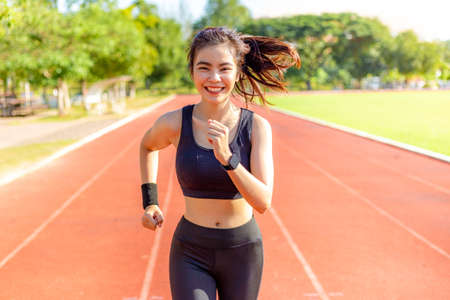 Beautiful happy young Asian woman running for her morning exercise at a running track with blurred track and trees in background, healthy lifestyleの写真素材