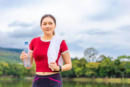 Beautiful Asian athlete woman drinking water during her break from morning  exercise at a lake park with view of lake and mountains in the background, healthy lifestyleの写真素材