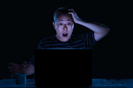 Middle age Asian male working on his computer at night in his home office, surprise to see content...