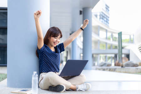 Happy young attractive asian business woman raises her arms up excitedly while looking at content on her laptop computer screen, sitting on the hallway floor of her office buildingの写真素材