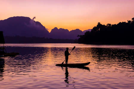 Man stand paddling on his kayak at Ratchaprapa dam, or known as Cheow Lan dam, with colorful sunrise backgroundの写真素材