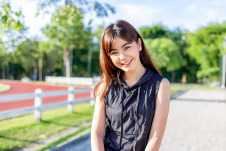 Happy beautiful young Asian woman smiling happily at camera during her exercise break in the morning at a running track with blurred trees and sky backgroundの写真素材