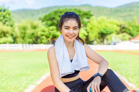 Happy beautiful young Asian woman sitting taking a rest after her morning exercise run at a running track surrounded by trees and mountains, good healthy lifestyle conceptの写真素材