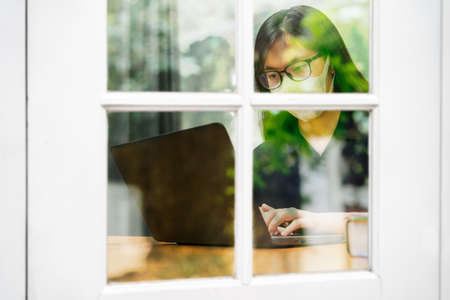 Young Asian woman wearing N95 mask and eyeglasses working on her computer at home, seen through window with reflection of trees, during city lockdown from COVID-19 virus, health and safety conceptの写真素材