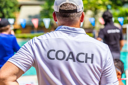 Male swimming coach in white COACH shirt standing in the sun watching his swimmers competing in the pool on a bright sunny dayの写真素材