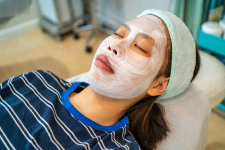 Young Asian female teenager using her smart phone while getting her acne and facial treatment with...