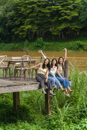 Three happy young Asian female friends sitting enjoying themselves on the wooden deck  by a river with big trees in background, friendship or young happy lifestyle conceptの写真素材