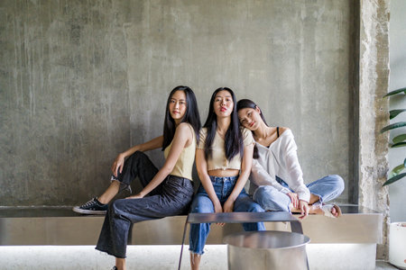 Three happy young Asian female friends casually pose sitting together inside a rustic room against bare concrete wall, young chic Asian modern lifestyle conceptの写真素材