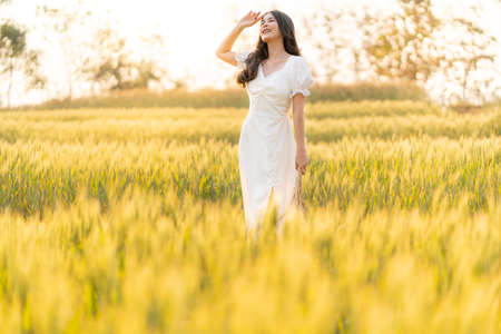 Happy young beautiful Asian woman in white dress standing in the barley field enjoying her time outdoor alone during sunset, lifestyle conceptの写真素材