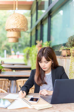 Happy young attractive business woman in her pink shirt writing down notes while working on her computer at outdoor patio of her office, working mobile or young entrepreneur conceptの写真素材