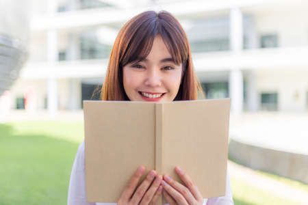 Happy young Asian woman smiling standing outside building and holding up a book in front of her face, education or lifestyle conceptの写真素材