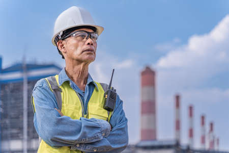 Senior engineer in his work cloths with white hard hat, safety glasses, and walkie talkie radio standing at the power plant site to supervise his projectの写真素材