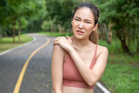 Young atractive Asian woman in fitness clothes putting her left hand over her pain right shoulder while standing at a running track of a local parkの写真素材