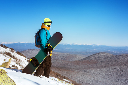 Snowboarder stands on the big rock keeps snowboard and looking far away. Sheregesh resort, Siberia, Russiaの写真素材