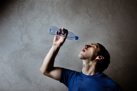 Muscle man drinking water from blue plastic bottle on gray wall backdropの写真素材
