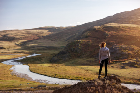 Lady stands on big rock against mountains and river at sunset time. Space for textの写真素材