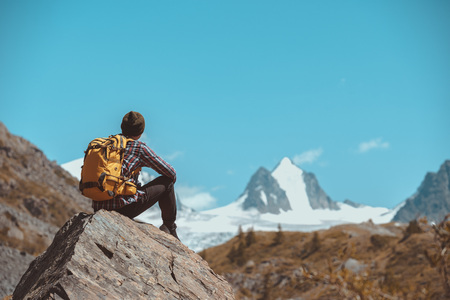 Man hiker with backpack sits on big rock and looks at white mountain with glacierの写真素材