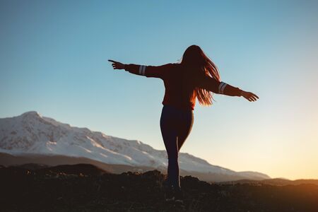 Beautiful slim girl walks with raised arms against sunset mountains and skyの写真素材