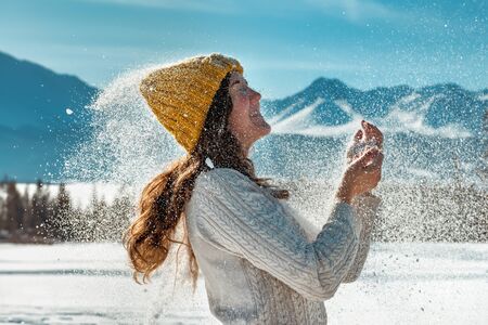 Happy beautiful girl plays with snow powder against forest and mountains