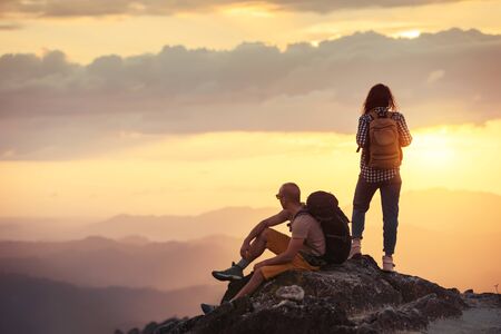 Couple hikers with backpacks are having rest on mountain top at sunset timeの写真素材