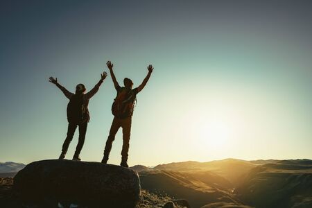 Two happy hikers stands with raised arms against sunset and enjoys mountains view