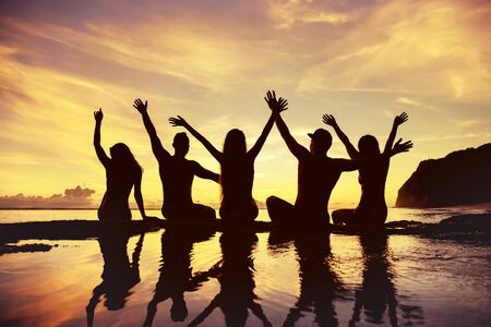 Five happy friends sits with raised arms against sunset sky and sea beachの写真素材