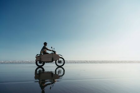 Real surfer with surfboard rides by enduro motorbike on the ocean beach. Bali island, Indonesia