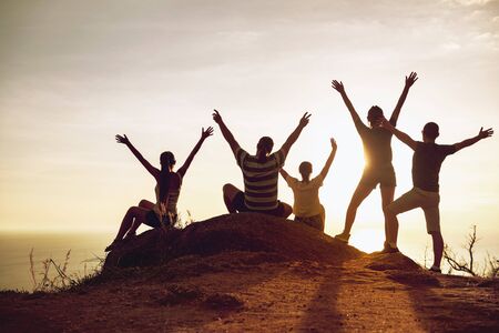 Group of happy tourists stands and sits with raised arms at sunset and having funの写真素材