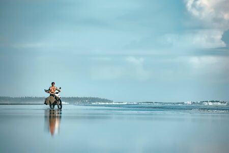 Authentic male surfer rides with surfboard on motorcycle at ocean beach. Real Bali surfing photoの写真素材