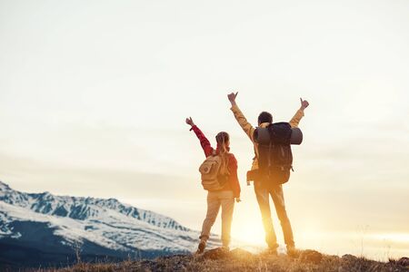 Two happy hikers with backpacks are standing with raised arms and looking at mountains and sunsetの写真素材