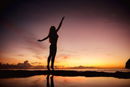 Happy woman in swimsuit stands with raised arms at sunset sea beachの写真素材