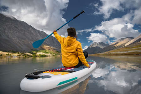 Stylish girl in yellow hoodie walks on sup board at the white mountain lake. Supboarding conceptの写真素材