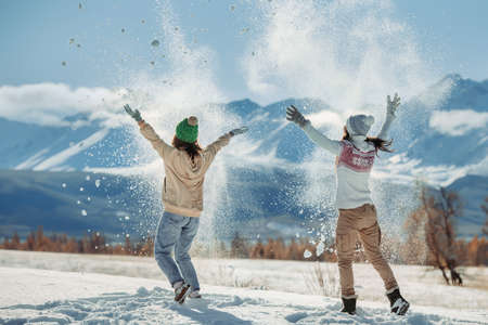 Two happy young girls are having fun and tossing snow in mountains. Winter holidays concept